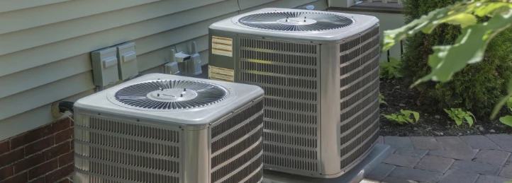 Two outdoor central air conditioning condenser units installed beside a home's exterior wall on a paved patio.