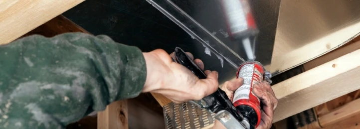 Technician applying sealant with a caulking gun to HVAC ductwork during residential system installation or repair.
